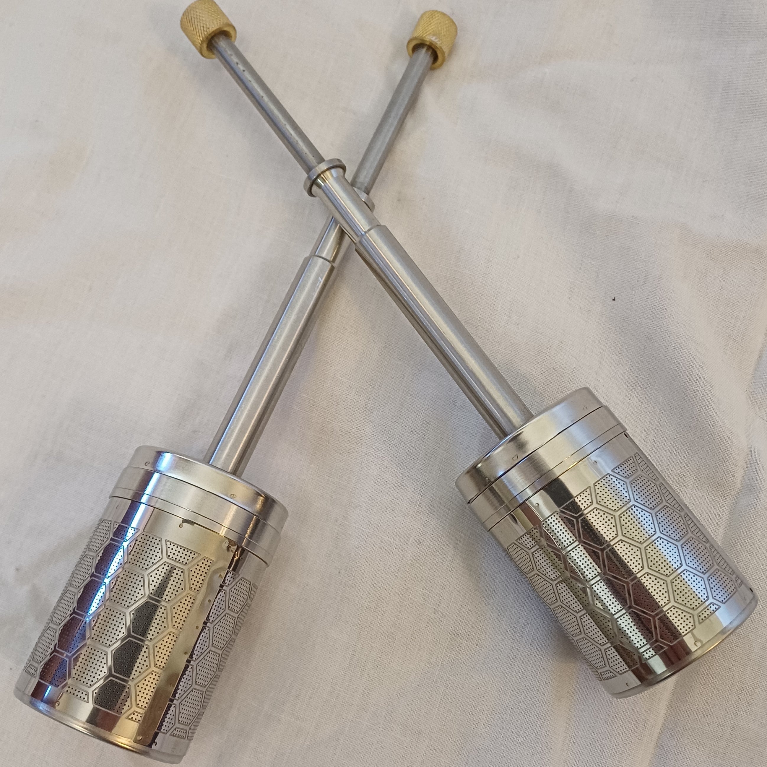Two silver metal tea infusers with textured design on a white fabric background